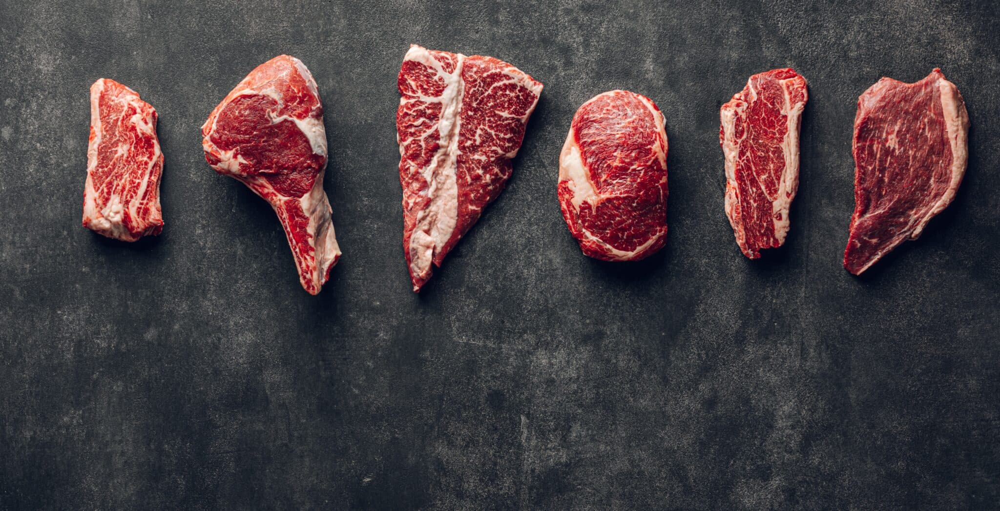 Variety of raw steak meat, laying on a dark coloured surface